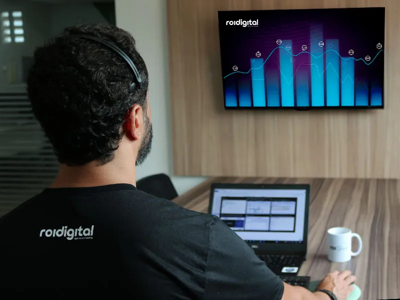 Profissional da ROI Digital de costas, trabalhando no notebook diante de uma TV com gráficos